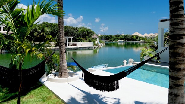Un área de piscina con una hamaca, palmeras y vistas a un lago con edificios al fondo.