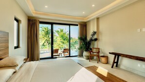 Un dormitorio amplio con una cama grande, un cabecero de madera, un balcón con vistas a palmeras y una zona de estar con un banco y sillones.