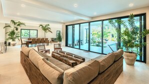 Amplia sala de estar con un sofá grande, mesa de comedor y sillas, rodeada de ventanas grandes y plantas en macetas.