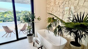 Un baño moderno con una bañera independiente, dos plantas en macetas y vistas a sillas exteriores a través de un ventanal.