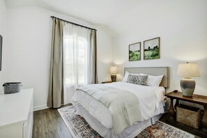 Un dormitorio con cama, mesitas de noche, lámpara, alfombra y ventana con cortinas.