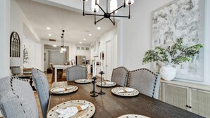 Comedor con una mesa de madera oscura dispuesta para seis personas, rodeada de sillas tapizadas con detalles abotonados. Un candelabro moderno cuelga arriba, y un gran cuadro abstracto adorna la pared.