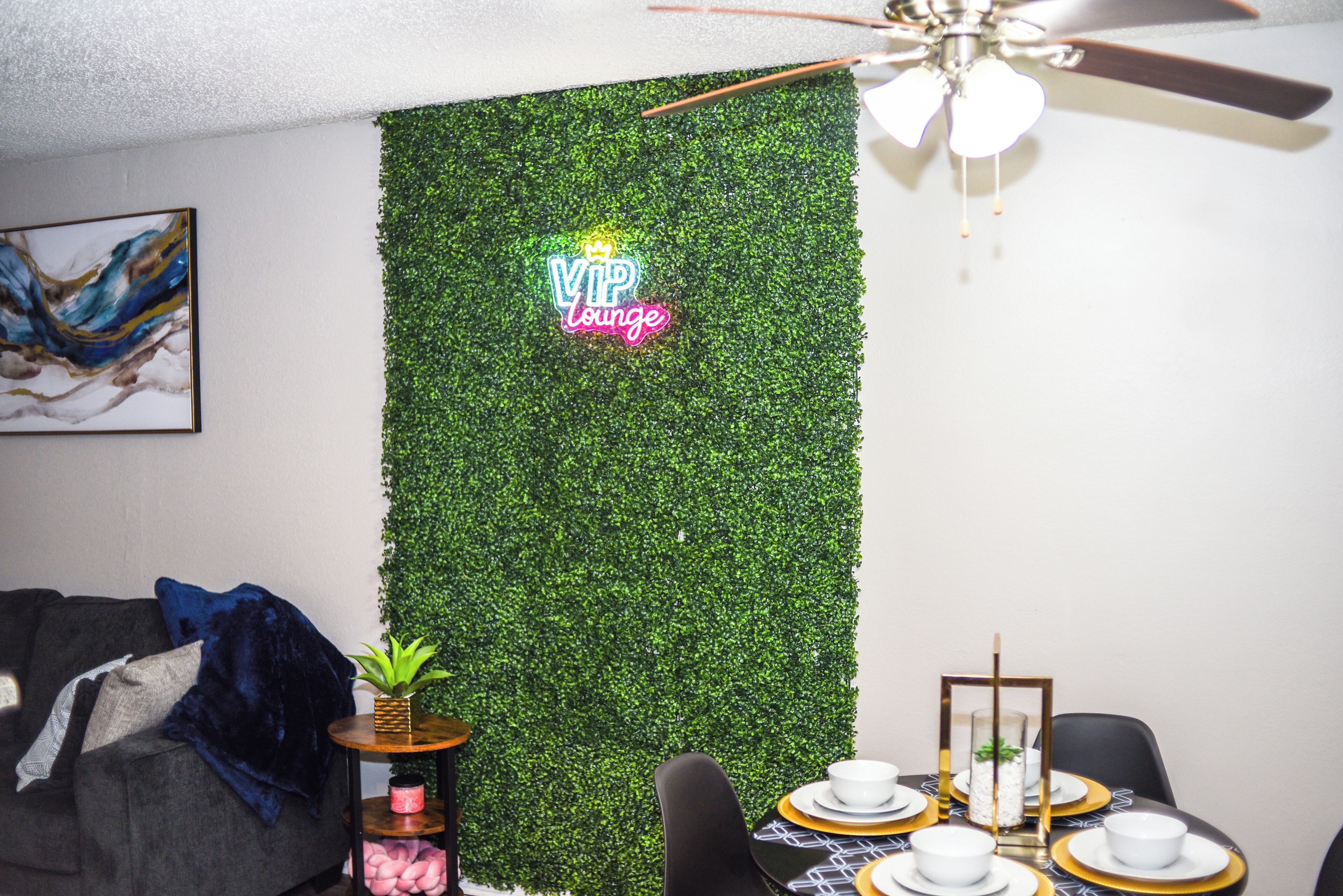 A room with a green wall, a ceiling fan, a dining table set with white plates and gold accents, and a small plant on a side table.