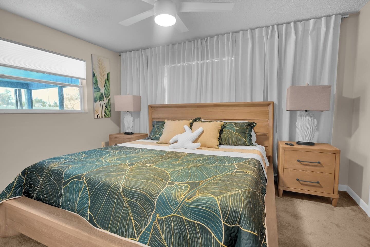 Un dormitorio con una cama grande, dos mesitas de noche, un ventilador de techo y una ventana con cortinas.