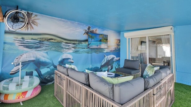 A room with a mural of dolphins, a fan, a sofa, and a table. - Pool + Ping Pong | Pets OK | Bonita Beach (Bonita Springs)