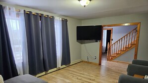 A living room with a wooden floor, grey sofas, a flat-screen TV mounted on the wall, and a staircase leading to another level. - Bayview Hideaway with a great mountains view and large peaceful property. (Whycocomagh)