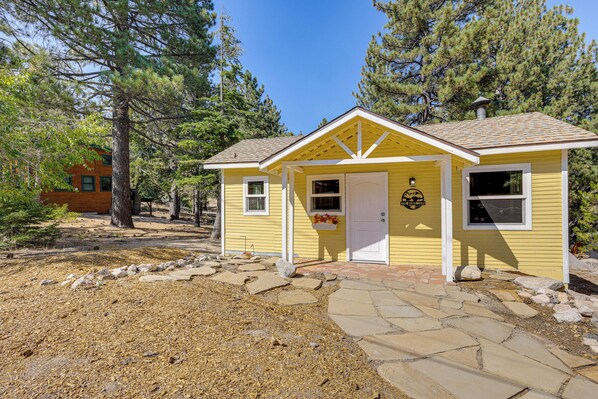 Une cabane jaune, avec un chemin de pierres menant à l’entrée, entourée de pins.