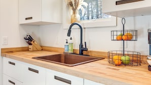 A kitchen with a wooden countertop, a sink, a window with a view of trees, a vase with dried flowers, and a fruit basket. - Private Hot Tub: Waterfront Home along Eagle River (Eagle River)
