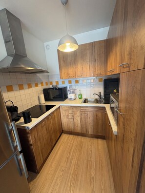 A kitchen with wooden cabinets, a stainless steel range hood, a microwave, a sink, and a hanging pendant light. - The Auryline (Thones)