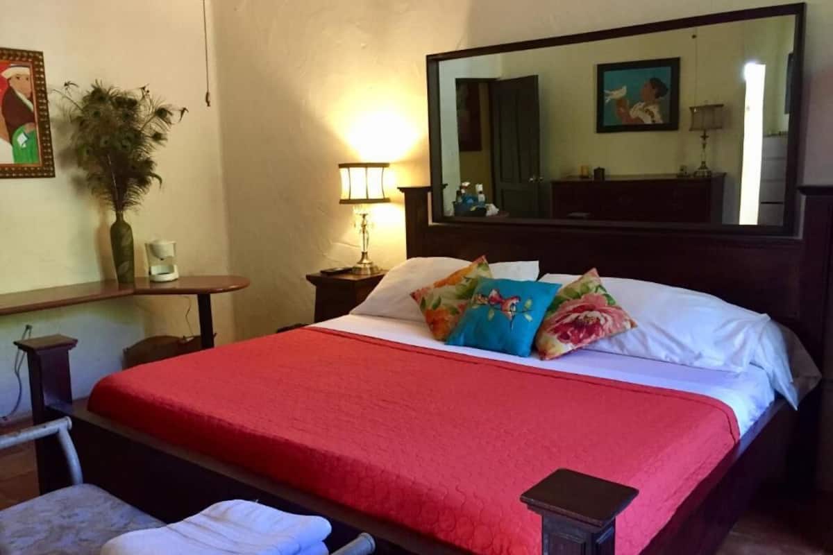 Habitación de hotel con una cama grande, una mesita de noche, una lámpara, un espejo y un cuadro colgado en la pared.