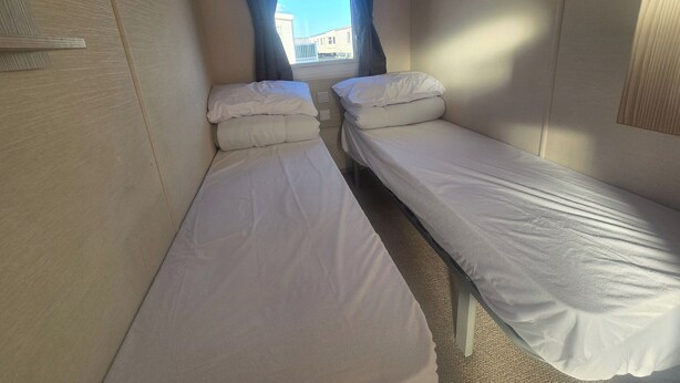 A small, narrow room with two single beds, a window with curtains, and a visible cityscape outside. - Collett 3 Bed Caravan Sleeps 6 With Wifi (Rhyl)