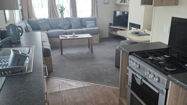 A modern kitchen with a stove, oven, and dining area. There is a sofa and a television in the living area. - Caravan Paul Sleeps 6 With Free Parking (Rhyl)