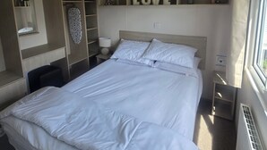 A modern hotel room with a large bed, built-in shelving with a 'LOVE' sign, a bedside table with a lamp, and a window with curtains. - Caravan Kirsty 3 Bedroom (Rhyl)