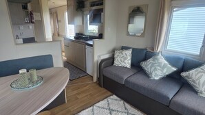 A compact RV interior with a kitchenette, a dining area, and a seating area with a sofa and a patterned rug. - Caravan Kirsty 3 Bedroom (Rhyl)