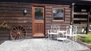 A rustic wooden building with a wooden door, two windows, and a white metal bench with a decorative backrest.