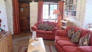 A cozy living room with a wooden table, two red sofas, a bookshelf, and a window with curtains. - Welsh Countryside Retreat - Felin Crewi Cottages (Machynlleth)