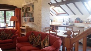 A cozy living room with a red sofa, a bookshelf, and a kitchen area with a skylight. - Welsh Countryside Retreat - Felin Crewi Cottages (Machynlleth)