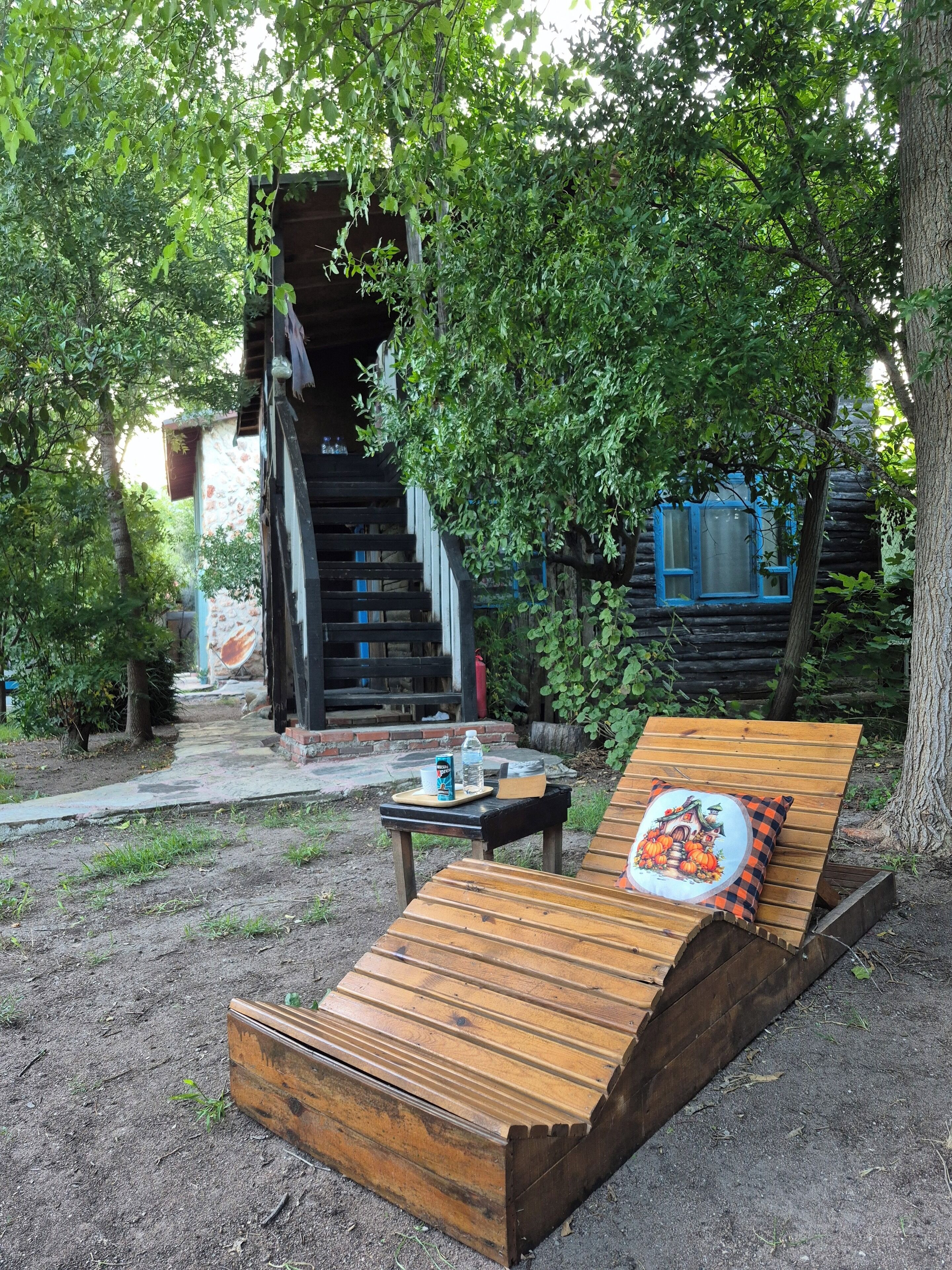 A wooden lounge chair with a cushion, a small table with a bottle and a can, and a staircase leading to a building with a blue window.