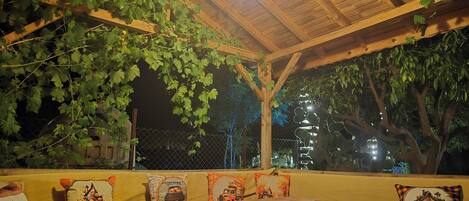 Un espacio de estar al aire libre cubierto, con bancos de madera y cojines, una mesa y una lámpara decorativa colgante.