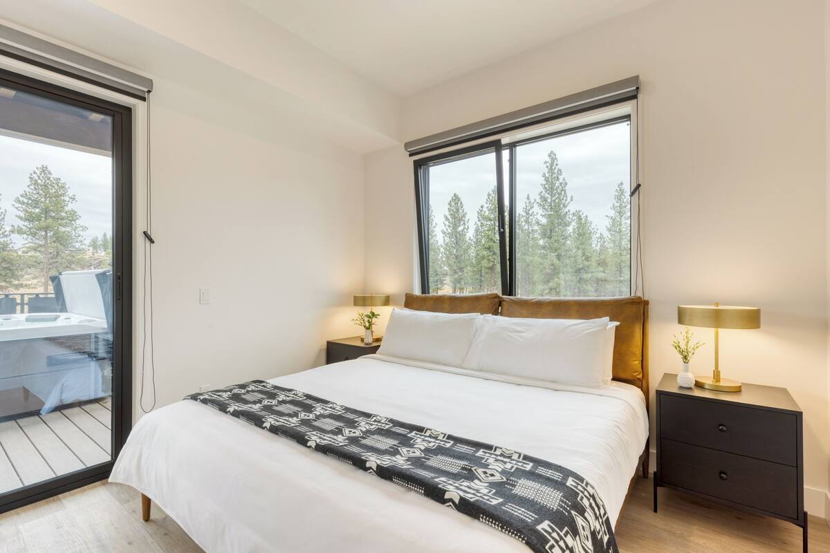 Een slaapkamer met een groot bed, twee nachtkastjes met lampen, een raam met uitzicht op bomen en een schuifdeur naar een balkon.