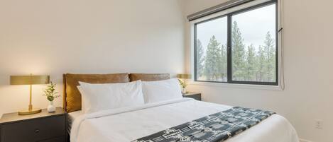 A bedroom with a large bed, two bedside tables with lamps, a window with a view of trees, and a white wall.
