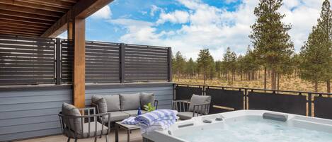 A wooden deck with a hot tub, seating area, and a view of trees and sky.