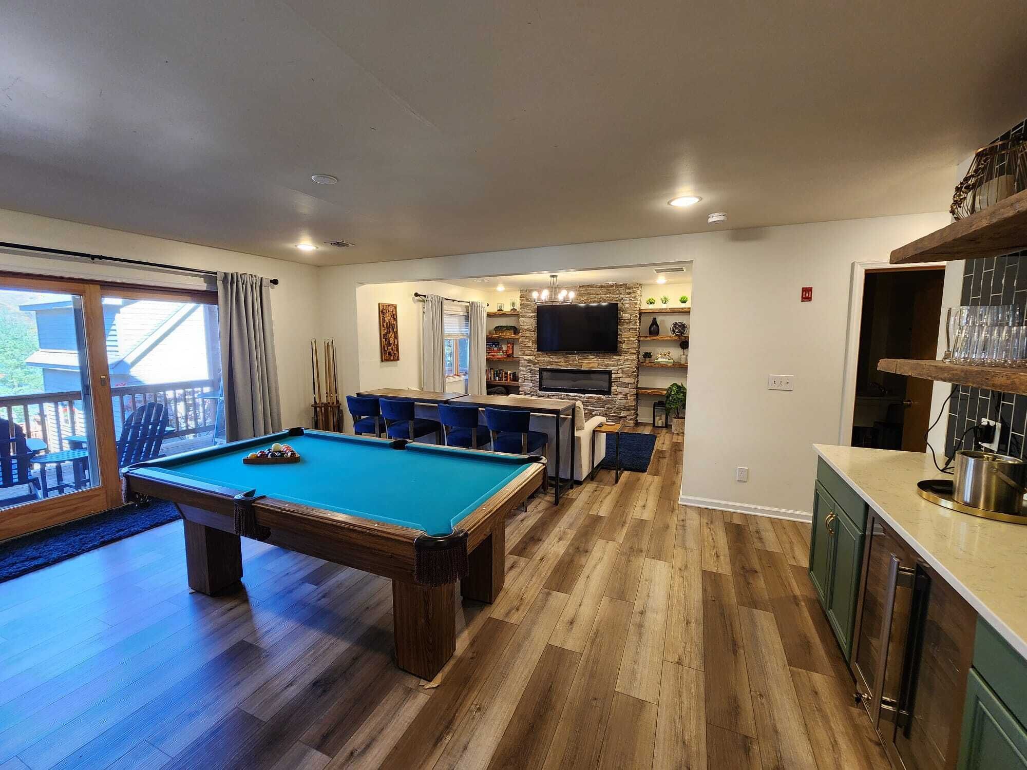 Une table de billard dans une pièce au plancher en bois, un coin repas avec des chaises bleues, une cuisine avec des meubles verts et un téléviseur fixé à un mur en pierre.
