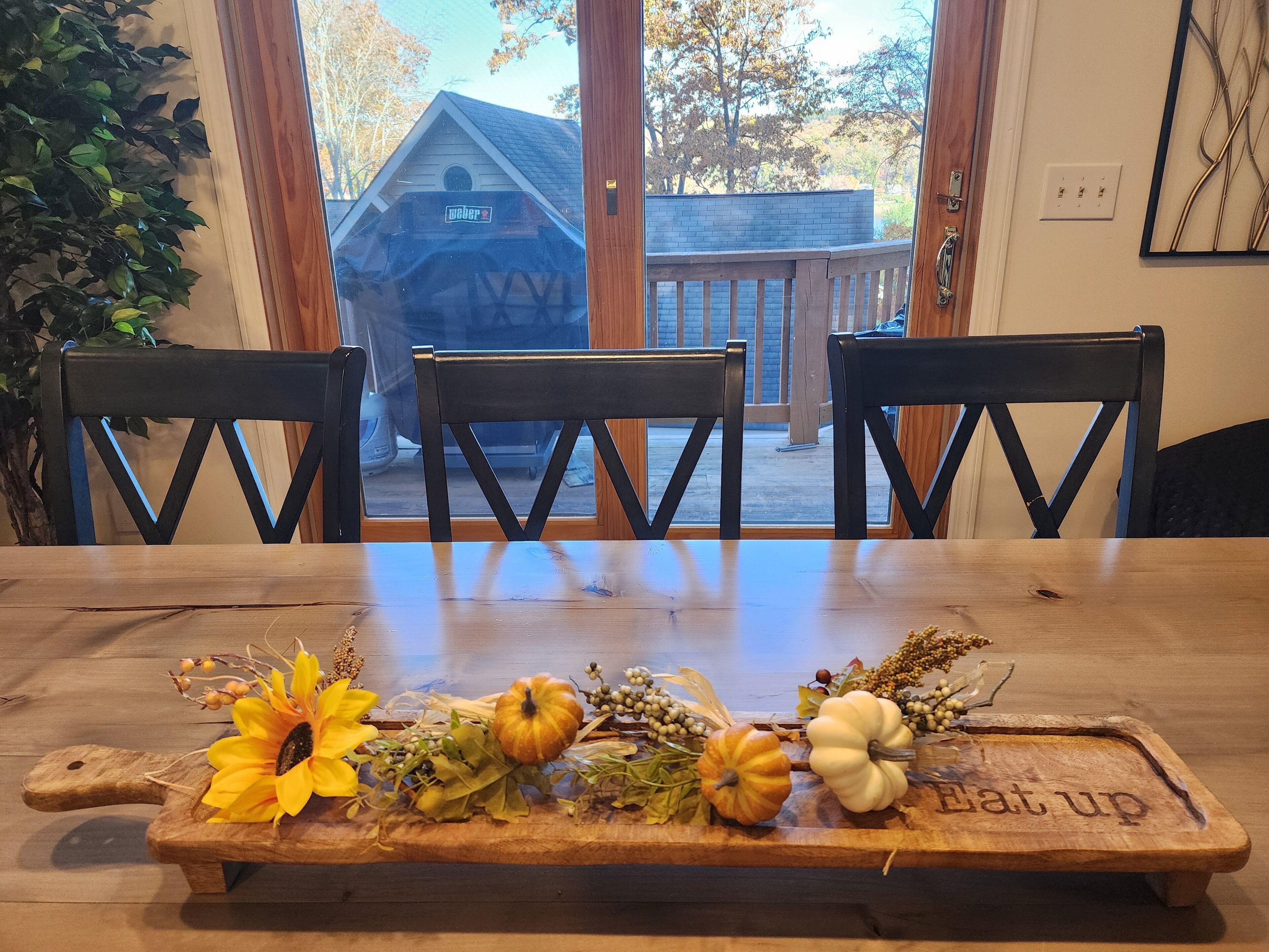 A dining table with chairs, a wooden tray with autumn decorations, and a view of a wooden building outside.