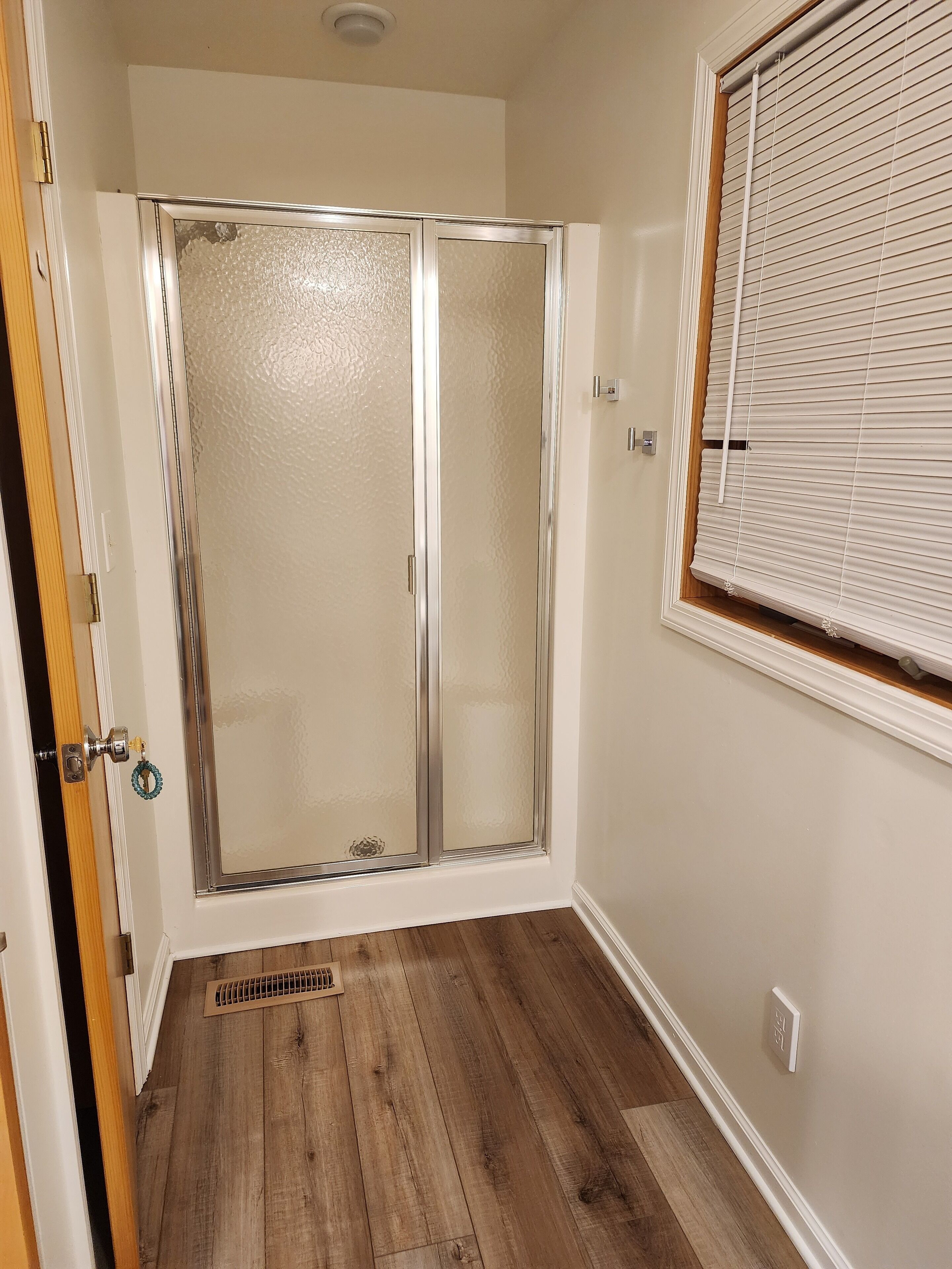 Ein Zimmer mit Glastür zu einem Duschbereich, einem Fenster mit Jalousien und Holzboden.