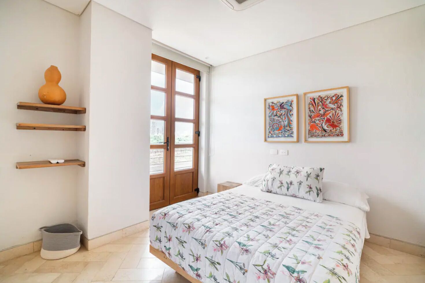 Een slaapkamer met een bed, een houten deur, een raam en twee kunstwerken in lijsten aan de muur.