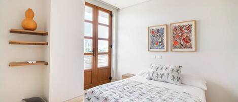 Een slaapkamer met een bed, een houten deur, een raam en twee kunstwerken in lijsten aan de muur.