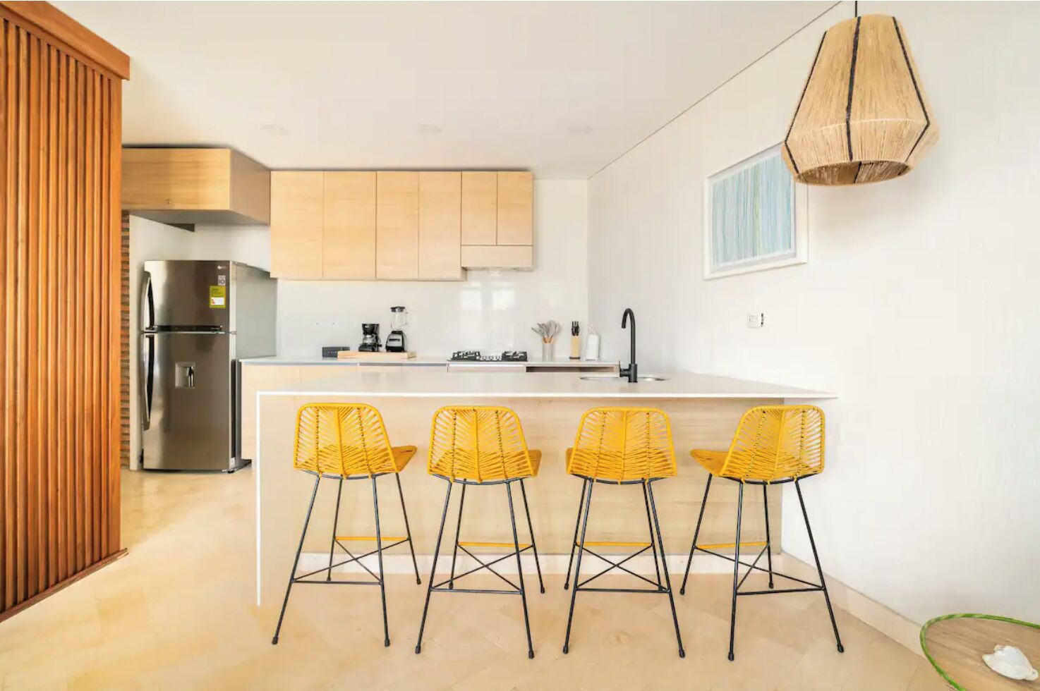 Een moderne keuken met een roestvrijstalen koelkast, houten kasten, een wit aanrecht en vier gele barkrukken.