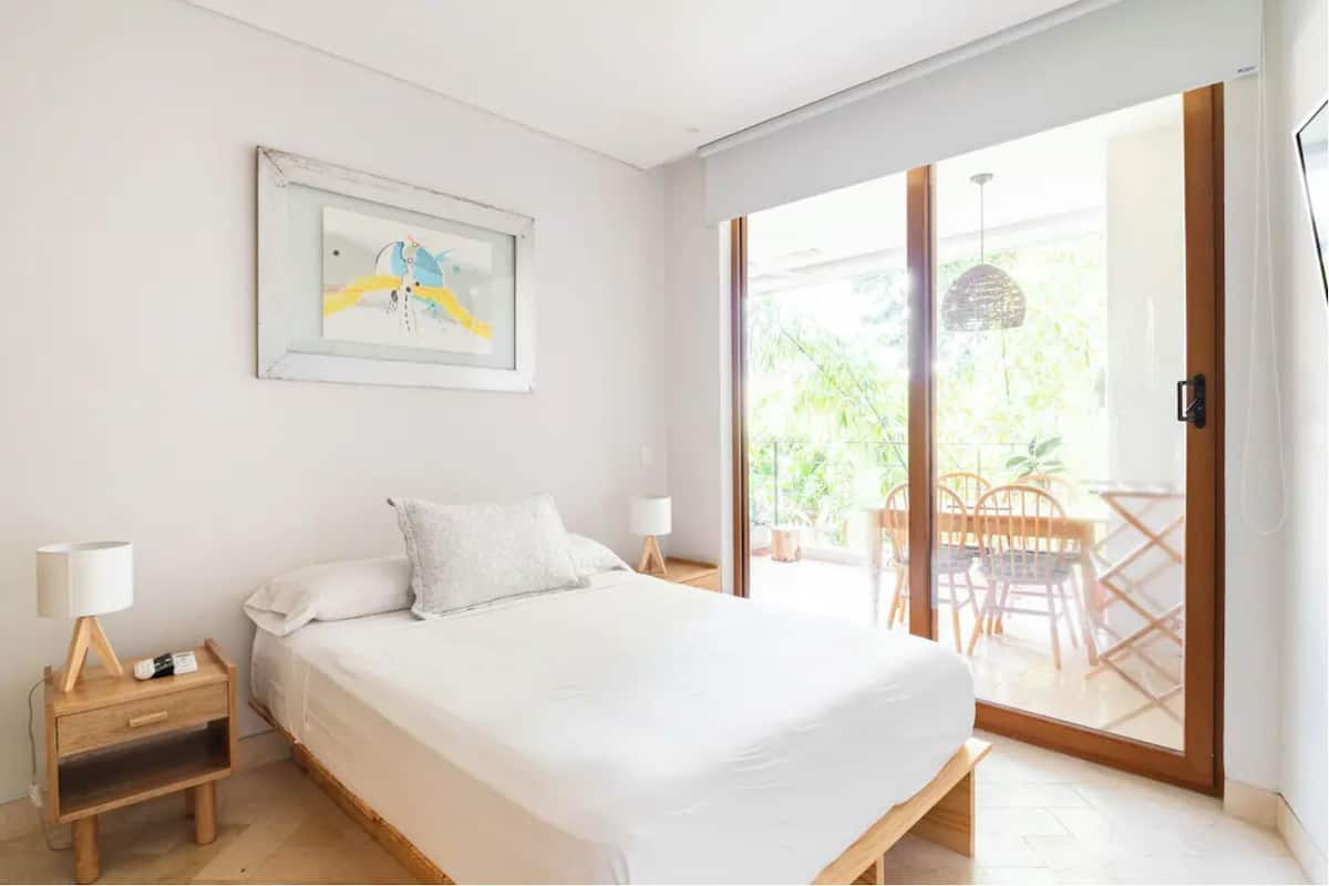 Un dormitorio moderno con una cama grande, una mesita de noche, una lámpara de noche y un ventanal corredero que da a un balcón con una mesa y sillas.