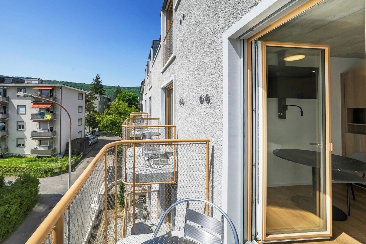A residential building with a balcony, a dining area with a table and chairs, and a view of neighboring buildings and greenery.