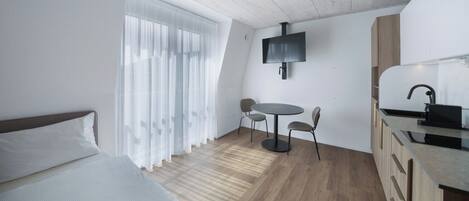 Una habitación compacta con cama, una pequeña mesa de comedor con sillas, un televisor montado en la pared y una kitchenette.