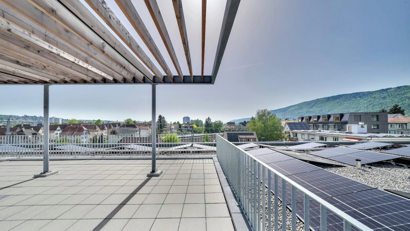 A rooftop terrace with solar panels, a pergola, and a view of a cityscape and hills.