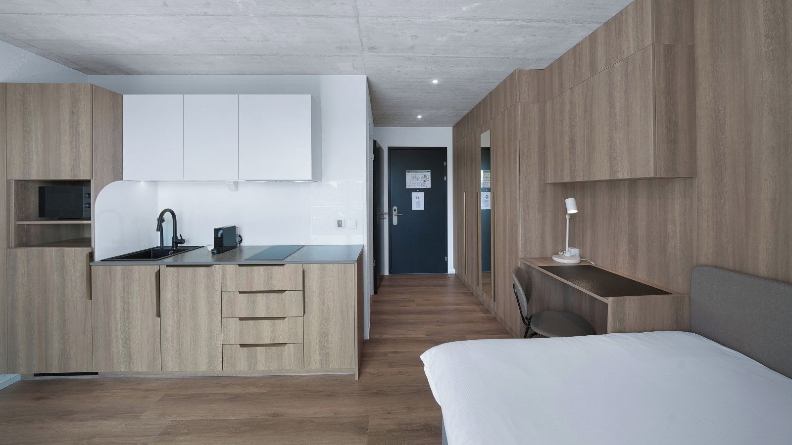 A modern hotel room with a kitchenette, a bed, a desk, and a chair.