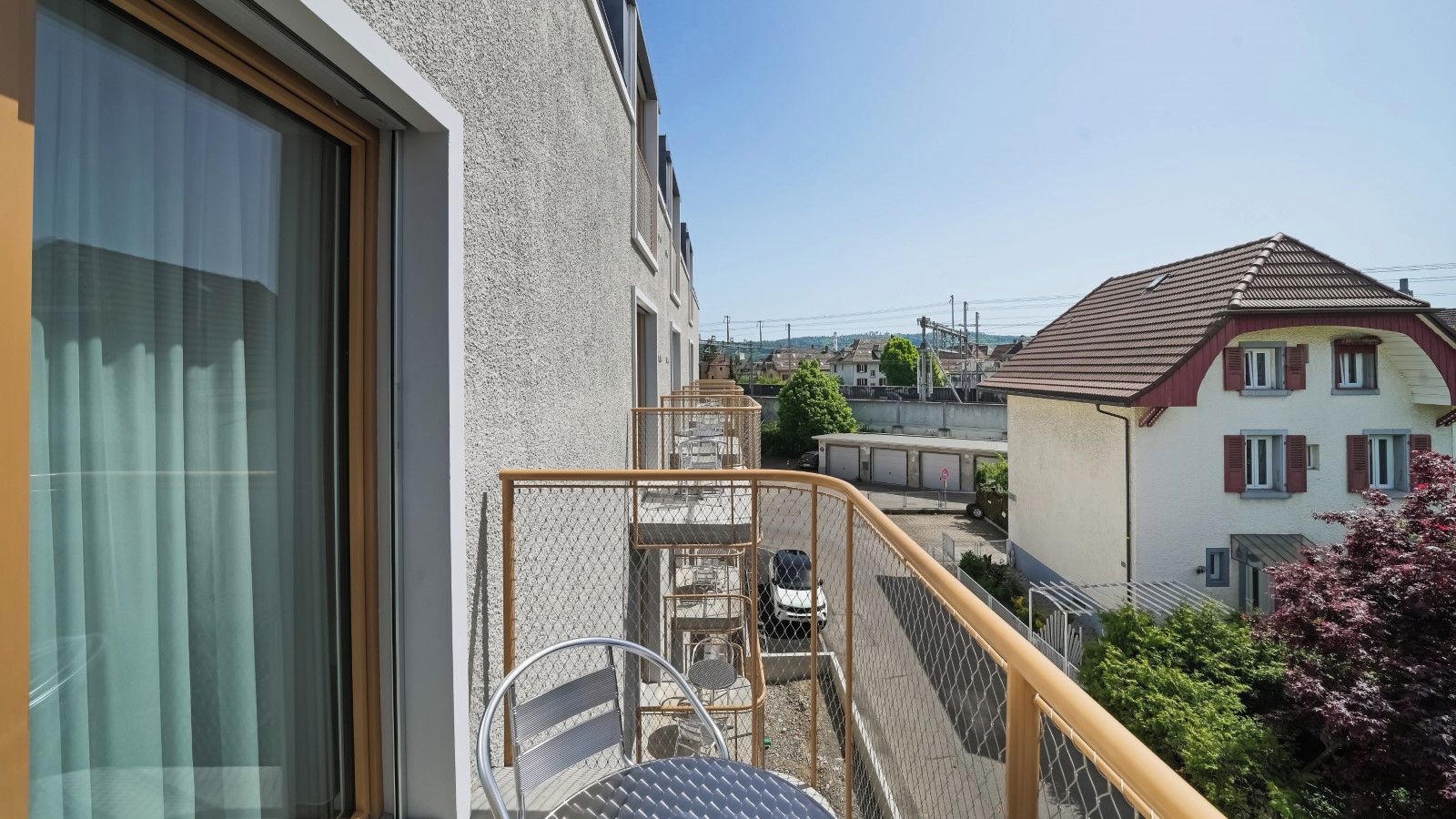 Un balcon avec une chaise en métal et une petite table, donnant sur un quartier résidentiel avec des maisons et un ciel dégagé.