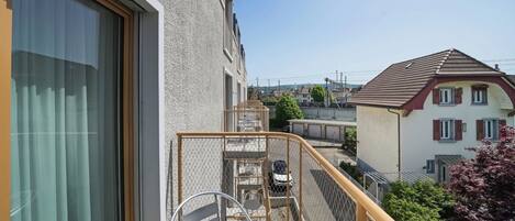 A balcony with a metal chair and a small table, overlooking a residential area with houses and a clear sky.