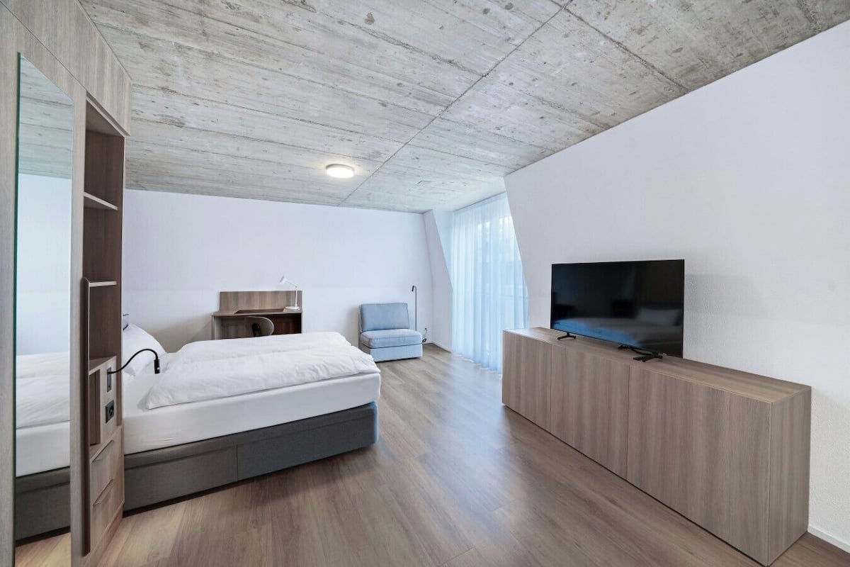 A modern hotel room with a wooden ceiling, a bed, a TV on a wooden stand, a desk with a chair, and a large window with sheer curtains.