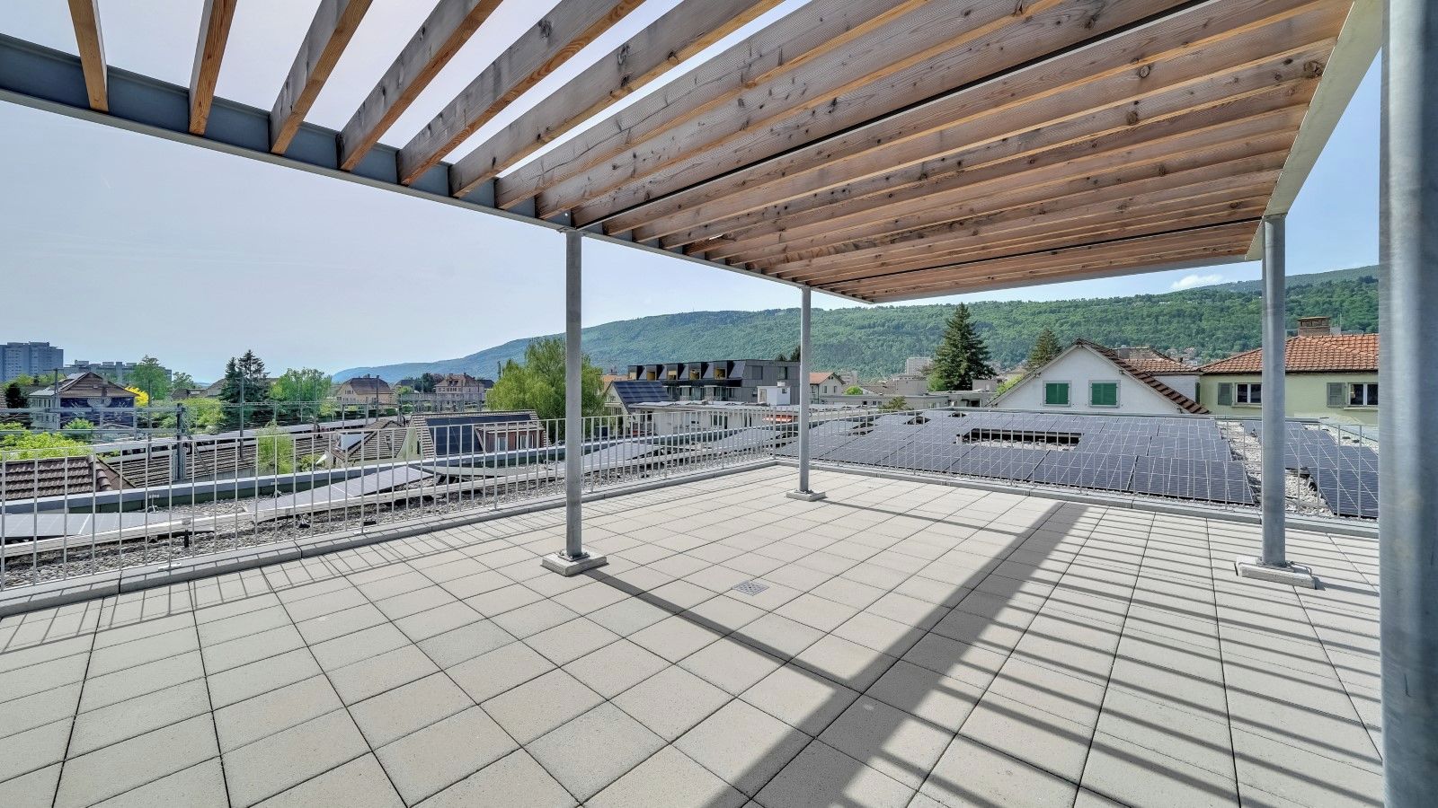 Eine Dachterrasse mit einer Holzpergola, Fliesenboden und Blick auf Gleise und umliegende Gebäude.