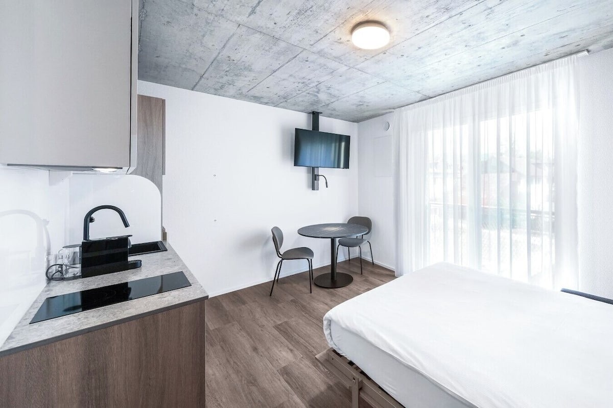 A modern hotel room with a bed, a small desk with a chair, a TV mounted on the wall, and a window with sheer curtains.
