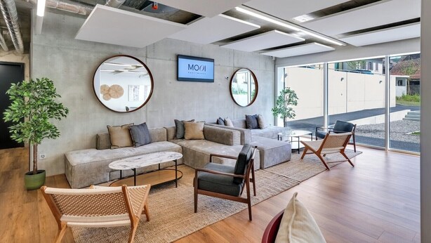 Un salon moderne avec un canapé d’angle, une table basse et deux fauteuils. On y trouve deux miroirs ronds accrochés aux murs, une plante en pot et une enseigne indiquant « MOOI Apartments ».