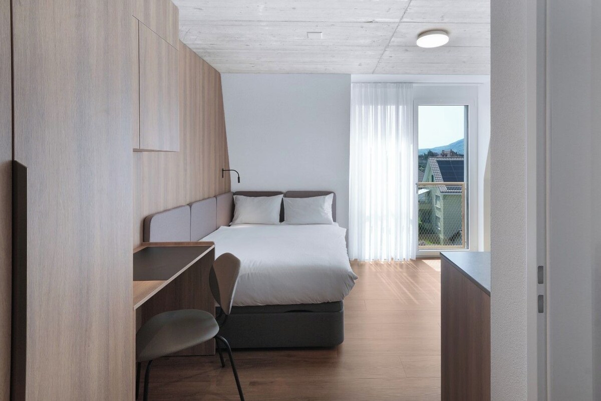 A modern bedroom with a bed, a desk, and a chair. There is a window with a view of buildings and a mountain.