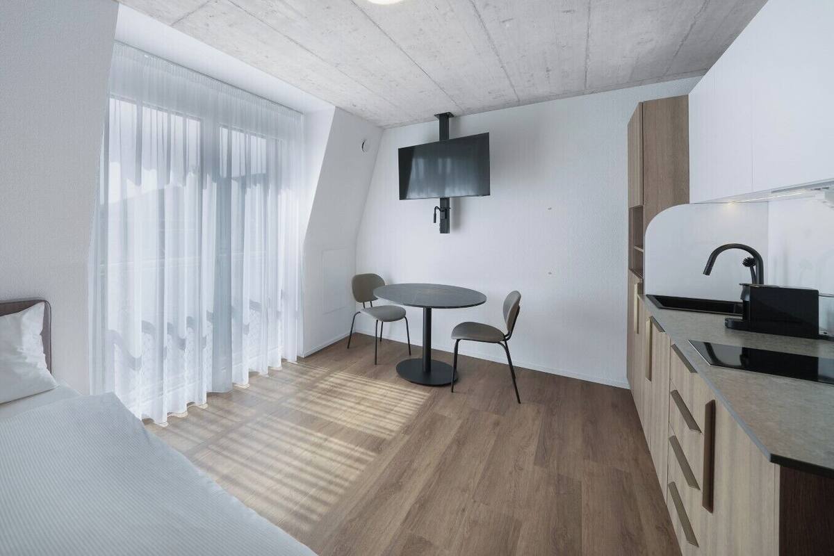 A compact room with a bed, a small dining table with chairs, a TV mounted on the wall, and a kitchenette area.