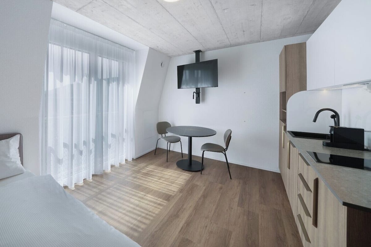 Une pièce de taille modeste comprenant un lit, une petite table à manger avec des chaises, un téléviseur fixé au mur et un coin cuisine.
