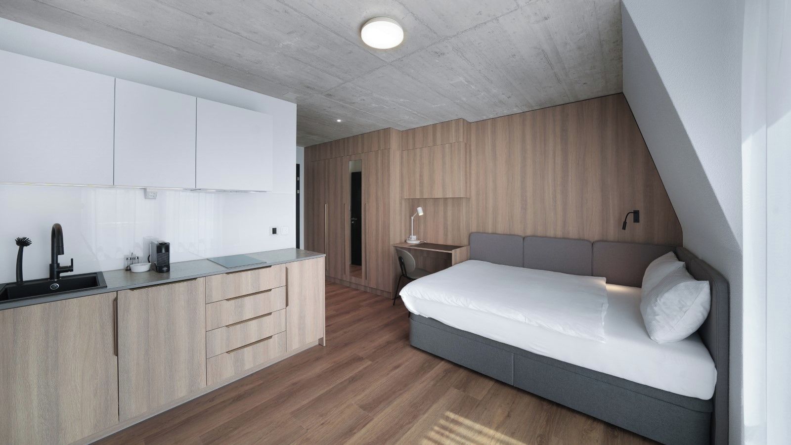 A modern bedroom with a built-in bed, a small desk, and a kitchenette.