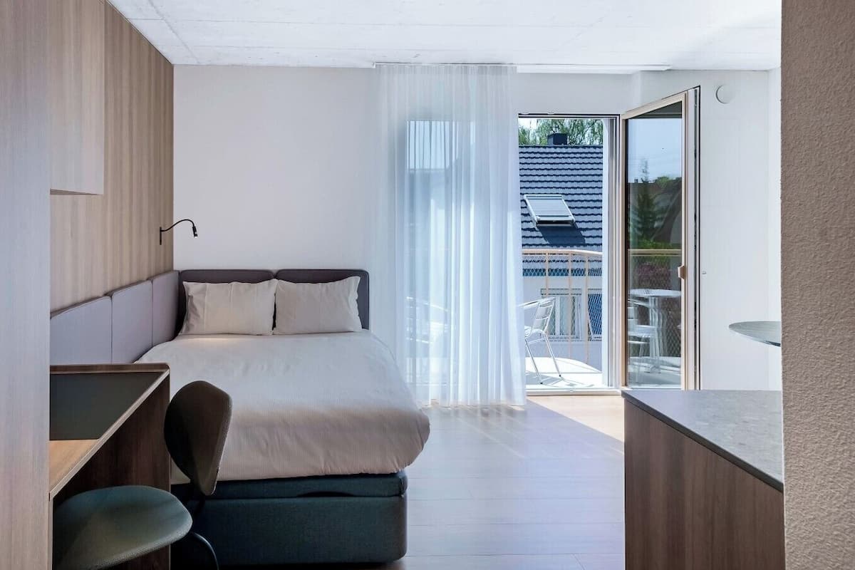 A modern hotel room with a bed, a desk, and a balcony with a view.
