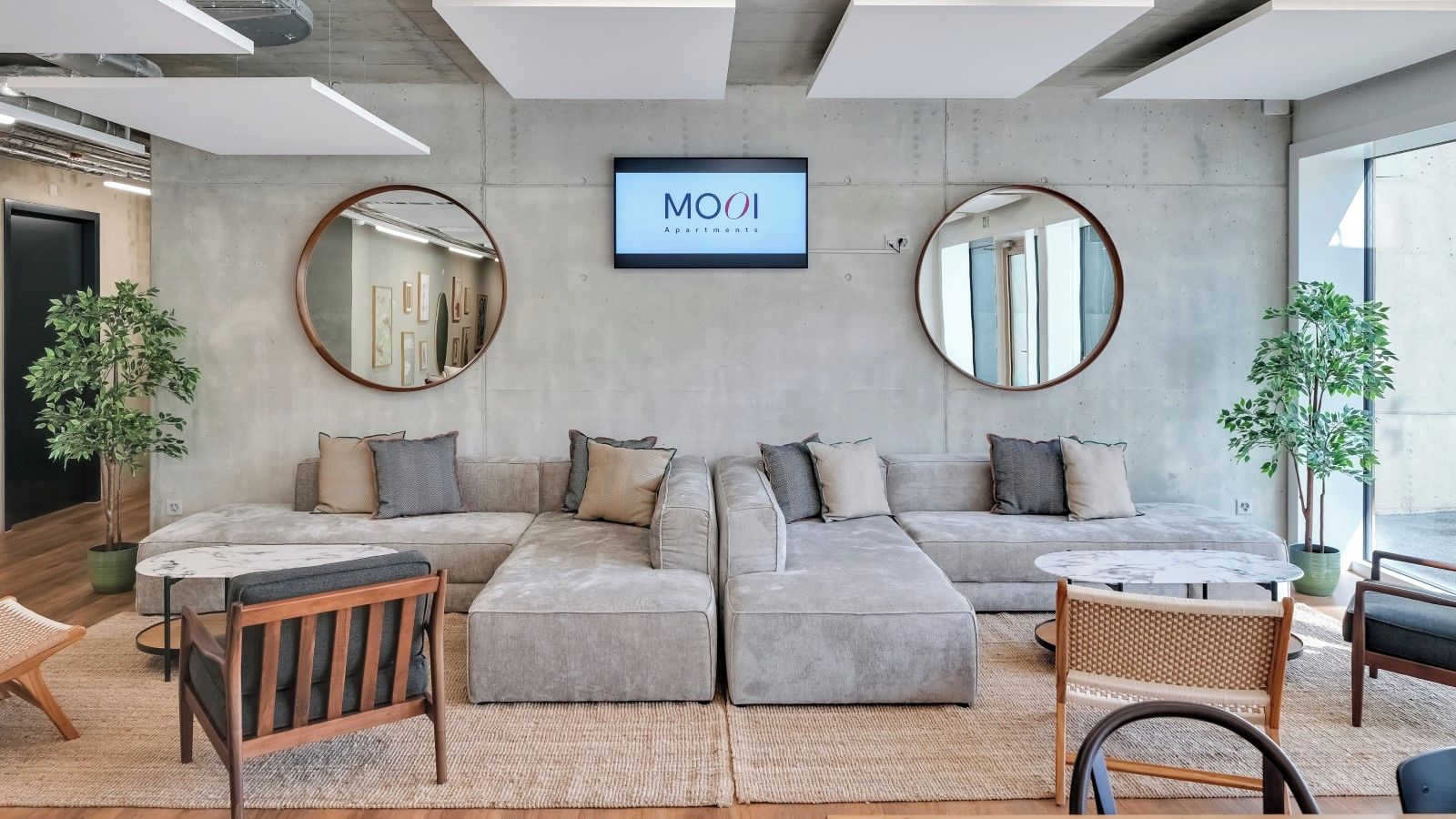 Ein modernes Wohnzimmer mit einem grauen Ecksofa, runden Spiegeln und einem Fernseher, der "MOOI Apartments" zeigt.