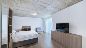 A modern hotel room with a wooden ceiling, a bed, a TV on a wooden stand, a desk with a chair, and a large window with sheer curtains. - Mooi Apartments Biel (Biel)